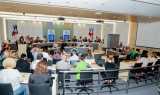 Large conference room with a panel of speakers at the front, French flags behind them, and attendees using laptops and notebooks.