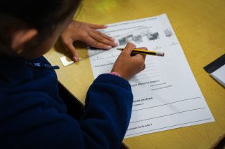 Child in a blue sweater writes on a French worksheet at a yellow desk, holding a pencil near the page.