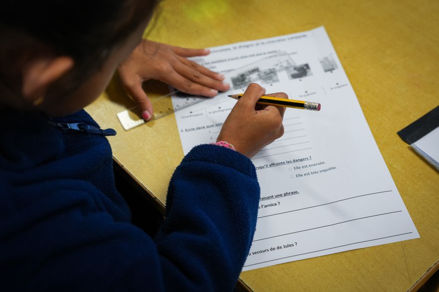 Child in a blue sweater writes on a French worksheet at a yellow desk, holding a pencil near the page.