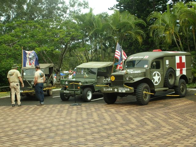 Green military ambulance with red cross parked on a paved area with flags and tropical trees in the background.
