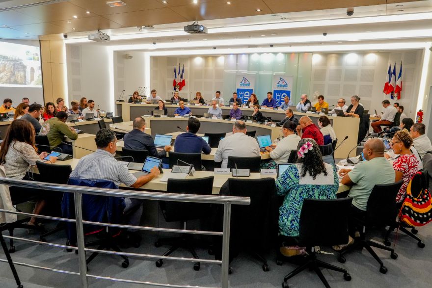 Panel of officials seated at a long desks in a conference room, with an audience using laptops and microphones; flags and banners in the background. Council meeting in progress.