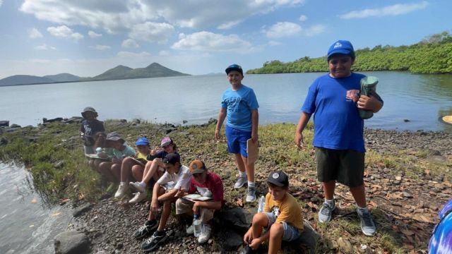 Group of kids by a rocky lakeshore, some sitting and writing/drawing, with calm water and hills in the distance.