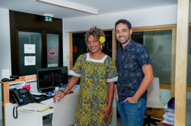 Two coworkers pose for a photo in an office reception area, the woman in a bright yellow patterned dress with a yellow flower in her hair and the man in a blue patterned shirt smiling beside her.