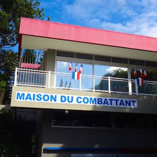 logo : Two-story building with a blue sign reading 'MAISON DU COMBATTANT' on the front, flags along the balcony, a pink awning, and a bright blue sky overhead.