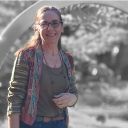 Smiling woman with glasses standing outdoors in a colorful cardigan and gray top, hands clasped, sunny park setting