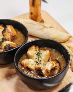 Two black bowls of creamy soup topped with croutons and herbs on a wooden board, with a torn baguette in the background.