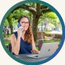 Woman seated at an outdoor cafe table, talking on a smartphone while using a silver laptop.