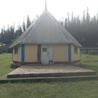 logo : Small yellow hut with a tall, gray metal roof and blue trim, sitting on a raised stone patio in a grassy area.