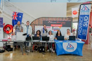 Group of six people standing and sitting behind a desk at an information booth, with banners reading Province Sud and Mont-Dore and various flags visible in the background (Australia flag on the left).