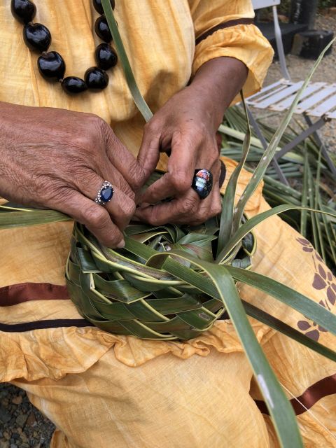 Close-up of elderly hands weaving a palm-leaf basket, with a blue gem ring and a beaded necklace over a yellow garment.