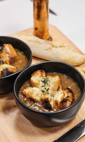 Two black bowls of creamy soup topped with croutons and herbs on a wooden board, with a torn baguette in the background.