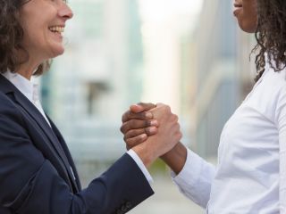 Two professionals in suits shake hands and smile, signaling agreement outdoors in a city setting.
