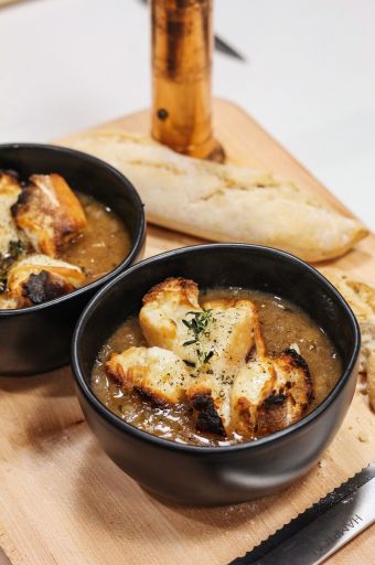 Two black bowls of creamy soup topped with croutons and herbs on a wooden board, with a torn baguette in the background.
