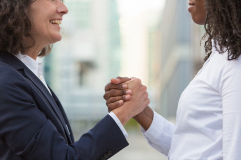 Two professionals in suits shake hands and smile, signaling agreement outdoors in a city setting.