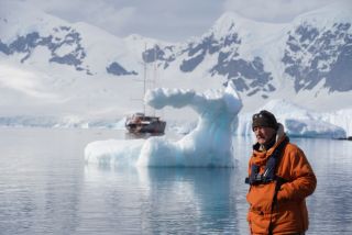 Man in an orange parka stands at a chilly shore with a blue iceberg arch and a sailing ship in the background.