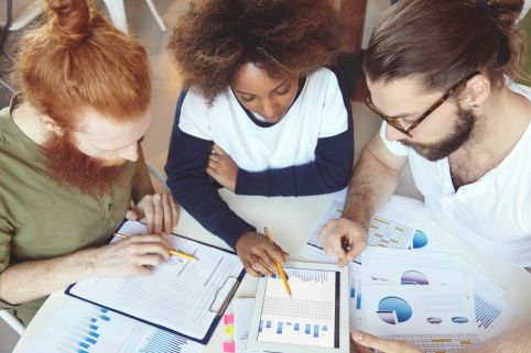 Three people collaborate at a table, examining charts, graphs, and a data-filled tablet during a group discussion.