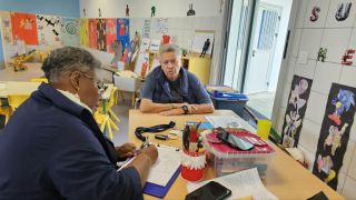 Two adults sit at a table in a bright art room, with colorful student artworks on the walls.