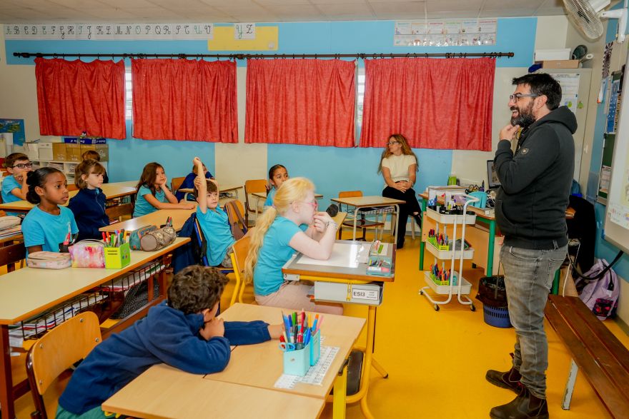 Bearded man in a dark hoodie speaks to a classroom full of young students seated at desks with blue walls and red curtains behind them.