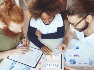 Three people collaborate at a table, examining charts, graphs, and a data-filled tablet during a group discussion.