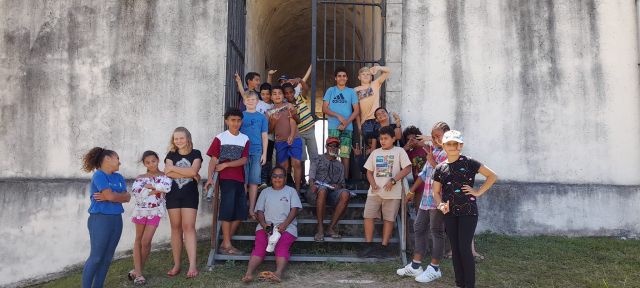 Group of kids and a few adults posing on a metal stair gate outside, smiling for a group photo on a sunny day.