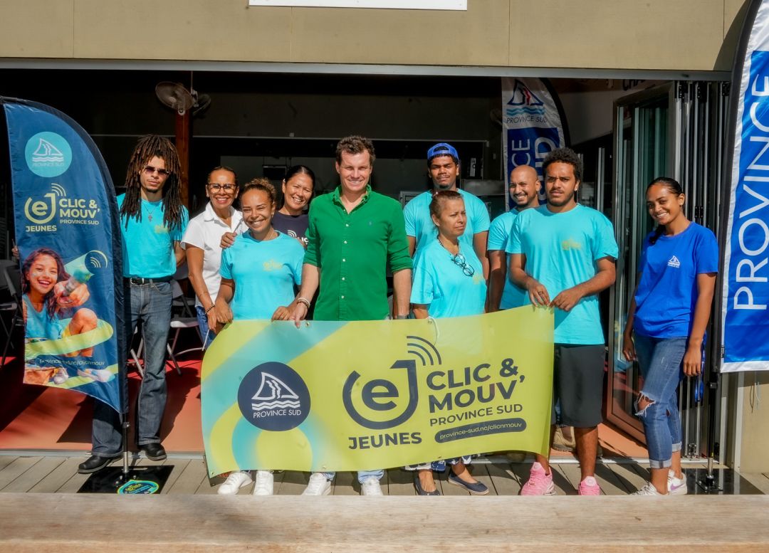 Group of diverse volunteers in turquoise shirts pose together behind a bright yellow banner at an outdoor event doorway, with Province Sud banners nearby.