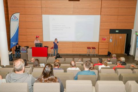 Speaker at a red lectern presenting to an audience in a conference hall, with a large projected slide on a wood-paneled wall behind them.