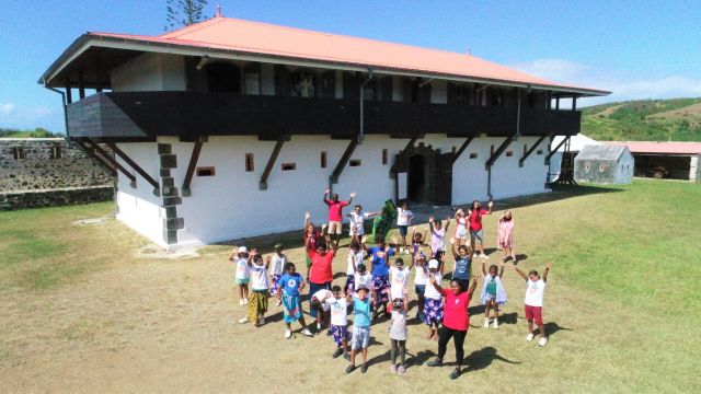 Group of children in colorful clothes waving at the camera in front of a two-story white building with a dark upper level and red roof, on a sunny day outside.