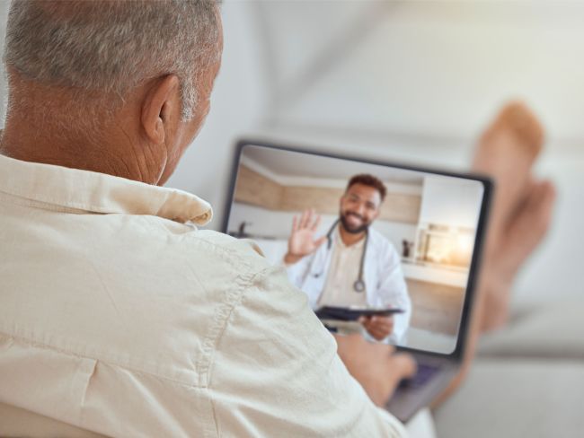 Older man shown from behind holding a tablet during a telemedicine video call, doctor waving on screen