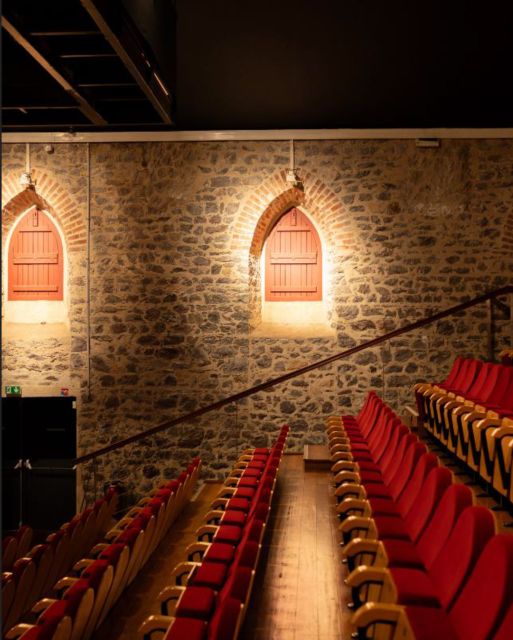 Empty theater with red and beige cushioned seats arranged in rows along a central aisle, stone walls and warm lighting on arched doorways.