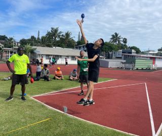 Two athletes leap to strike a blue ball during a track-side sport drill while teammates watch on a sunny day.