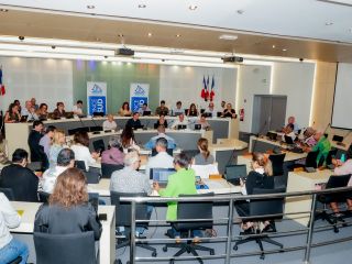 Large conference room with a panel of speakers at the front, French flags behind them, and attendees using laptops and notebooks.
