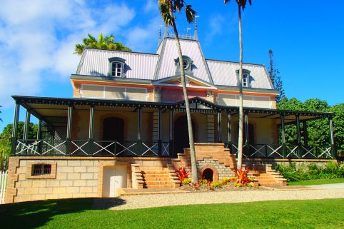 Two-story house with a metal roof, brick steps, and a wraparound veranda under a bright blue sky with palm trees in front.