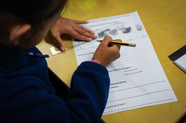 Child in a blue sweater writes on a French worksheet at a yellow desk, holding a pencil near the page.