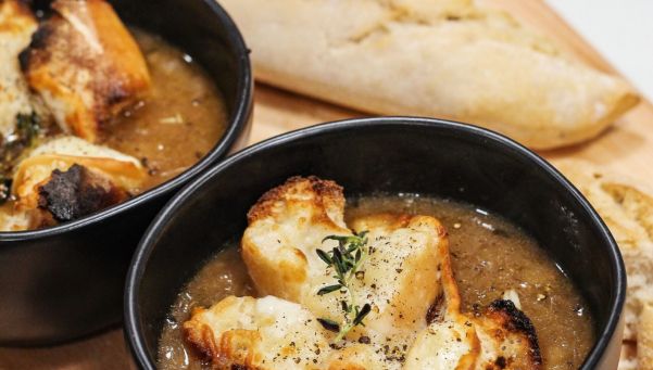 Two black bowls of creamy soup topped with croutons and herbs on a wooden board, with a torn baguette in the background.