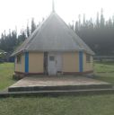 Small yellow hut with a tall, gray metal roof and blue trim, sitting on a raised stone patio in a grassy area.