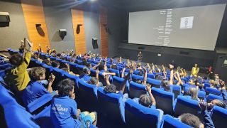 Large lecture hall filled with students in blue uniforms raising hands as a speaker presents at the front.