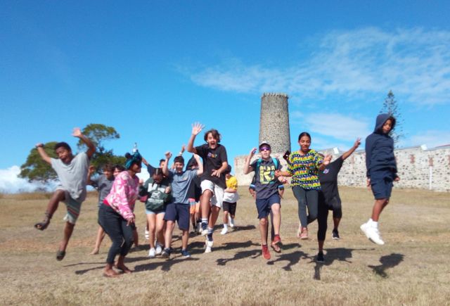 Group of children jumping and running in a dry field, with a stone tower and wall in the background under a bright blue sky.