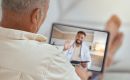 Older man shown from behind holding a tablet during a telemedicine video call, doctor waving on screen