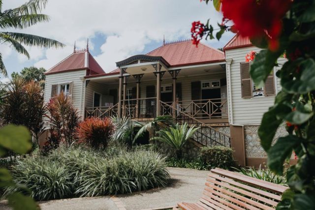 Historic house with a red roof, wraparound porch, and tropical garden in front, with a wooden bench in the foreground.