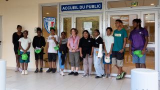 Group of teens and a woman posing outside a police station entrance, holding green caps. The door and a 'Accueil du Public' sign are visible behind them.