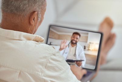 Older man shown from behind holding a tablet during a telemedicine video call, doctor waving on screen