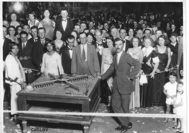 Large crowd in formal attire gathered around a tabletop model ship display at a social event, with people posing toward the camera.