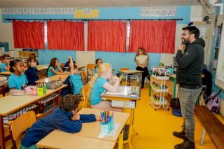 Bearded man in a dark hoodie speaks to a classroom full of young students seated at desks with blue walls and red curtains behind them.